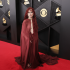 A woman with long, curly red hair and a sheer, flowing maroon gown stands on a red carpet with a black and gold patterned wall behind her.